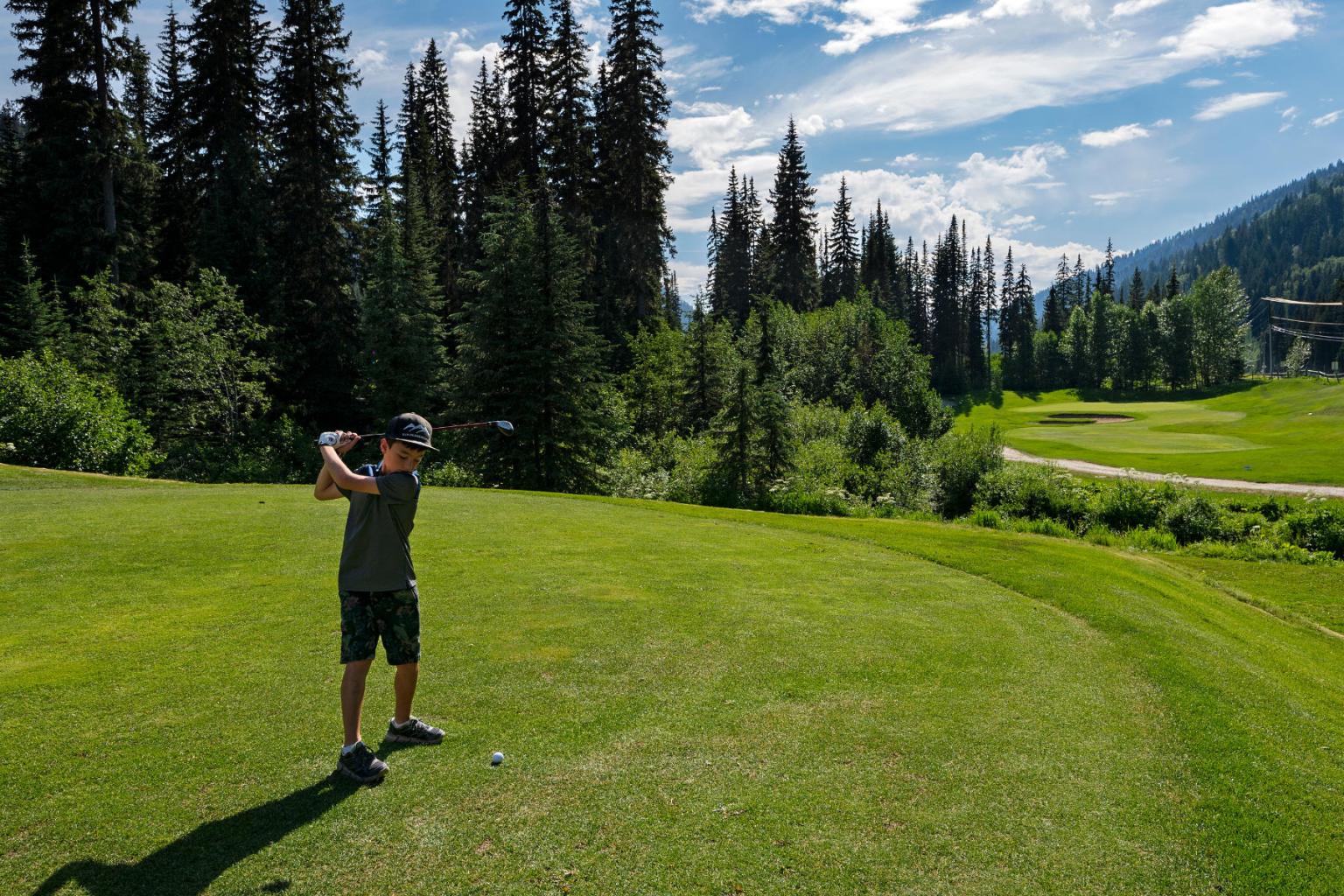 Young boy hitting a golf ball in the sun with trees surrounding him 