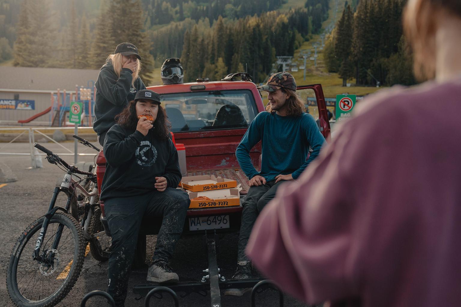 A woman in a pink shirt approaches a group of three mountain bikers tailgating on a red pick up truck with pizza boxes.