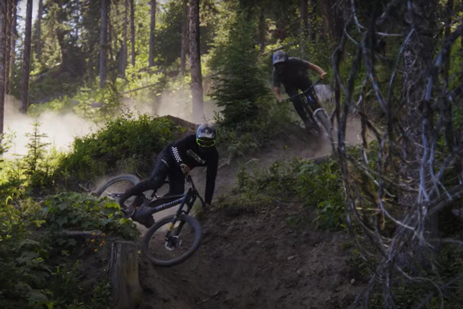Two downhill bikers riding down a trail in the forest with trees around them