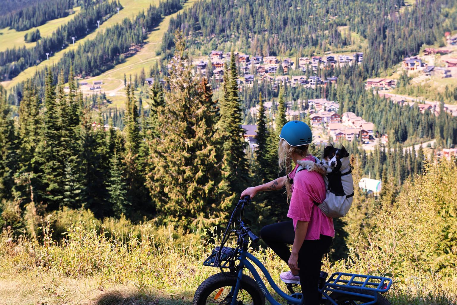 Woman riding bike with dog in backpack and sun peaks village in background