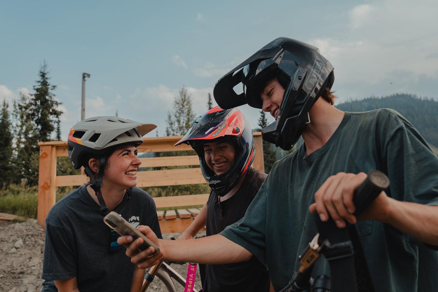Three mountain bike riders stand in front of a wooden feature and look at an iPhone while smiling.