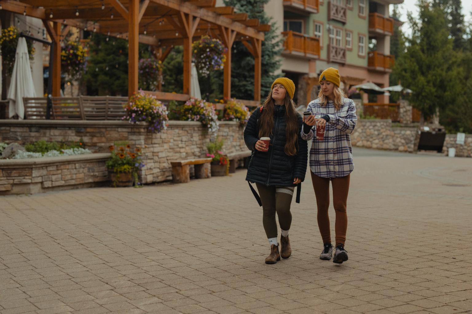 Two women walking through the village drinking coffee 