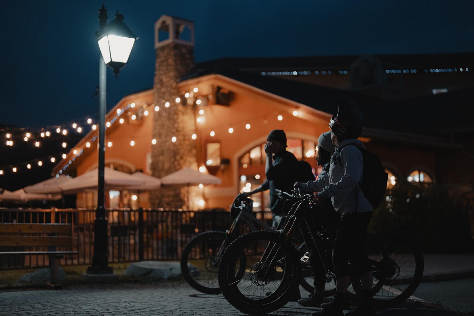 Three people walk their bikes at night past a patio glowing with outdoor string lights.