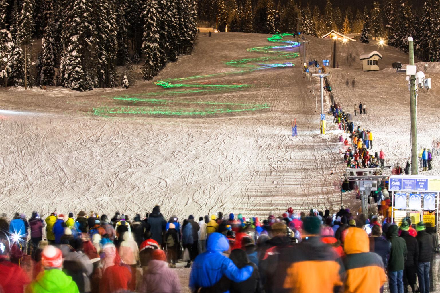People standing at the base of the village carpet with kids skiing down the run with green glowsticks