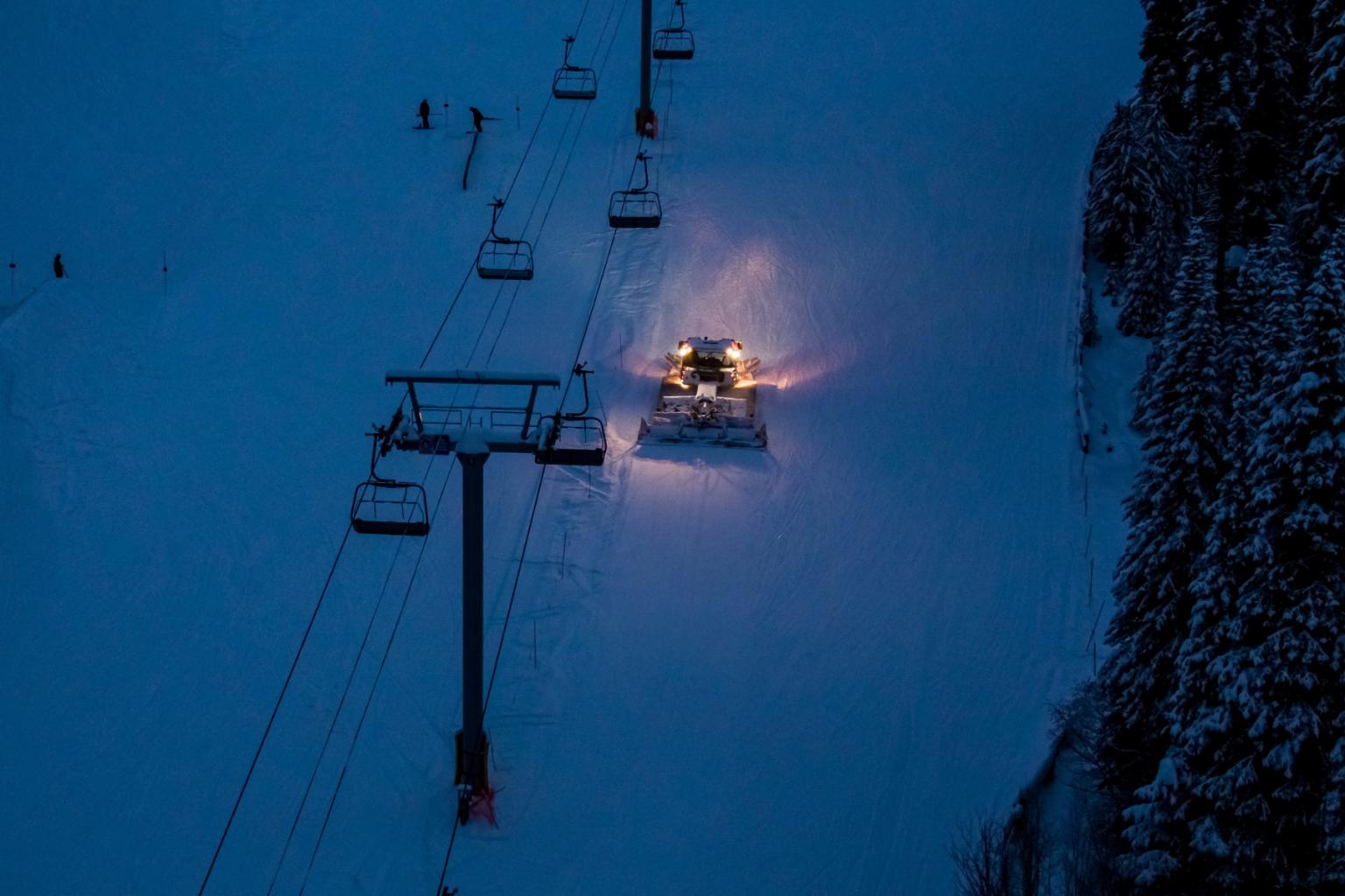 Aerial image of a groomer driving on the snow under a chairlift at night. 