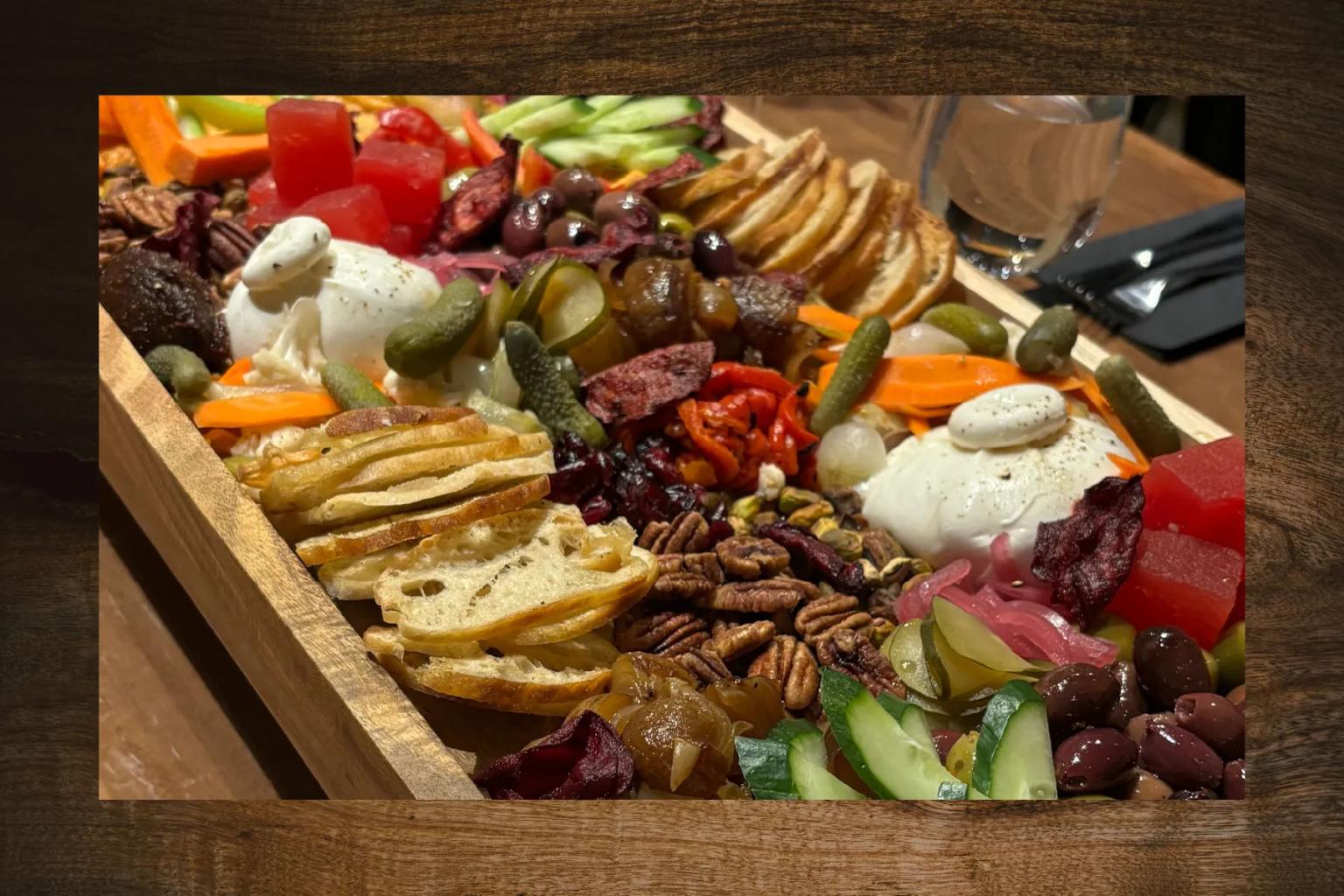 A charcuterie board with thin slices of baguette, burrata cheese, olives, cucumbers, walnuts, and quince jelly, accompanied by a glass of water and cutlery.