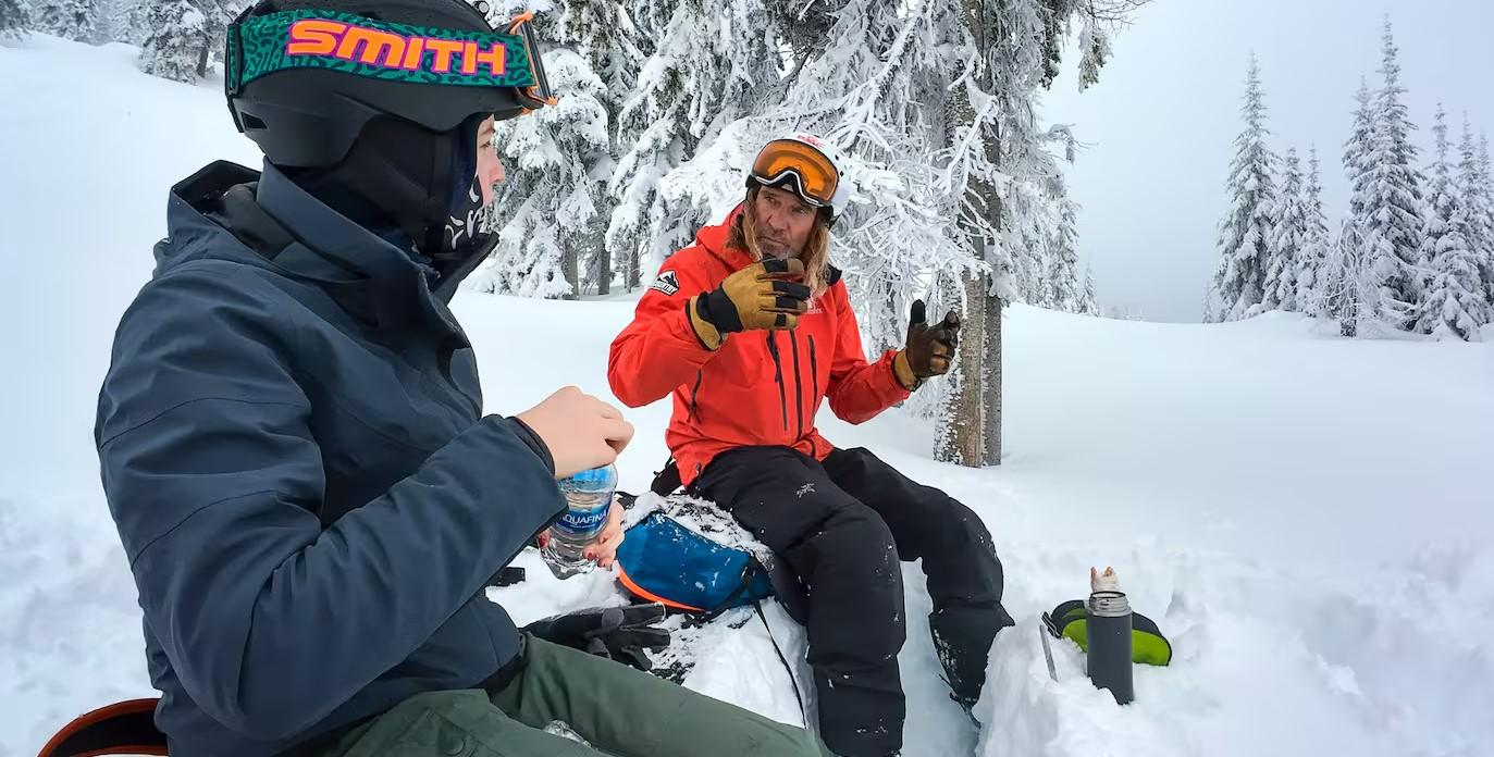 Young Skier sitting with Guide learning about tree wells eating lunch in powder