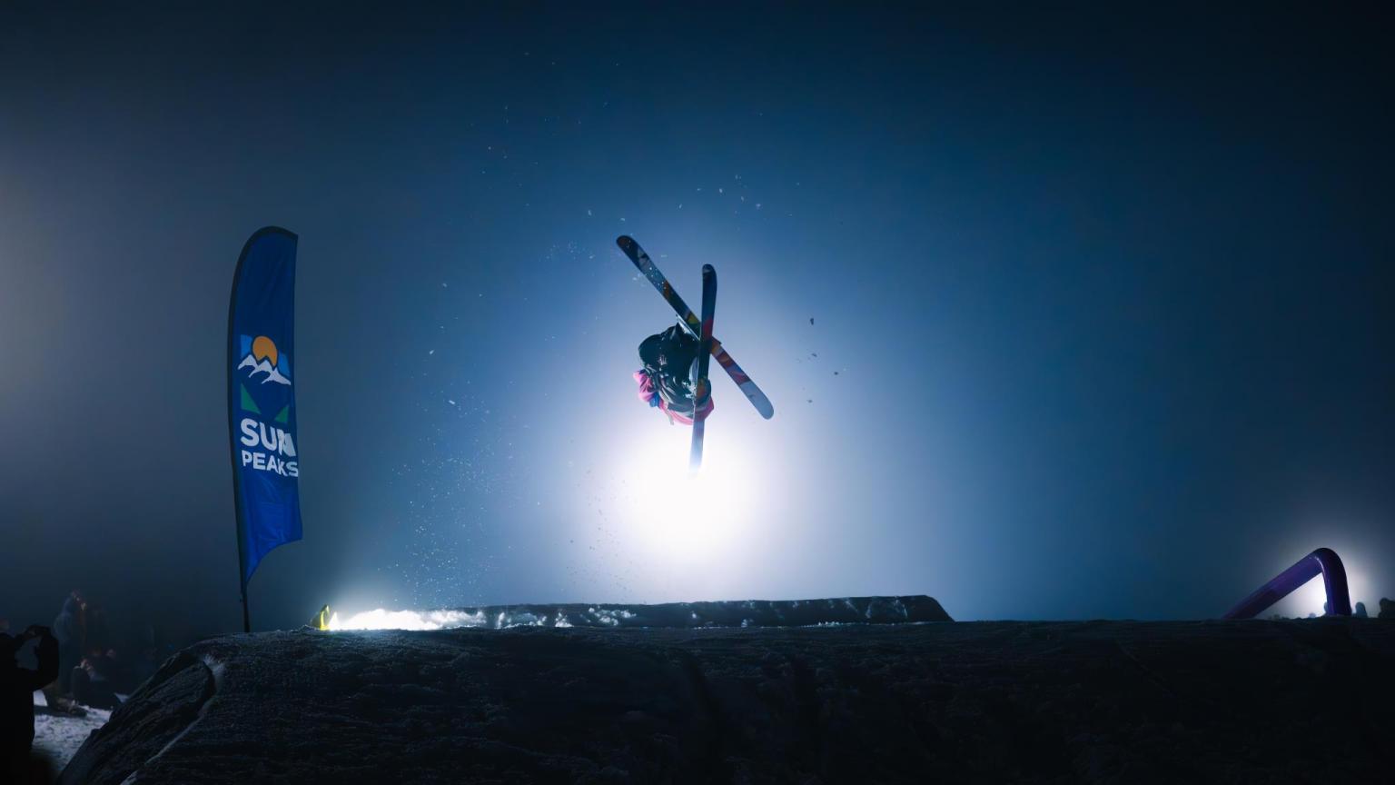 Skier doing a flip while hitting a jump with a light flashing behind them