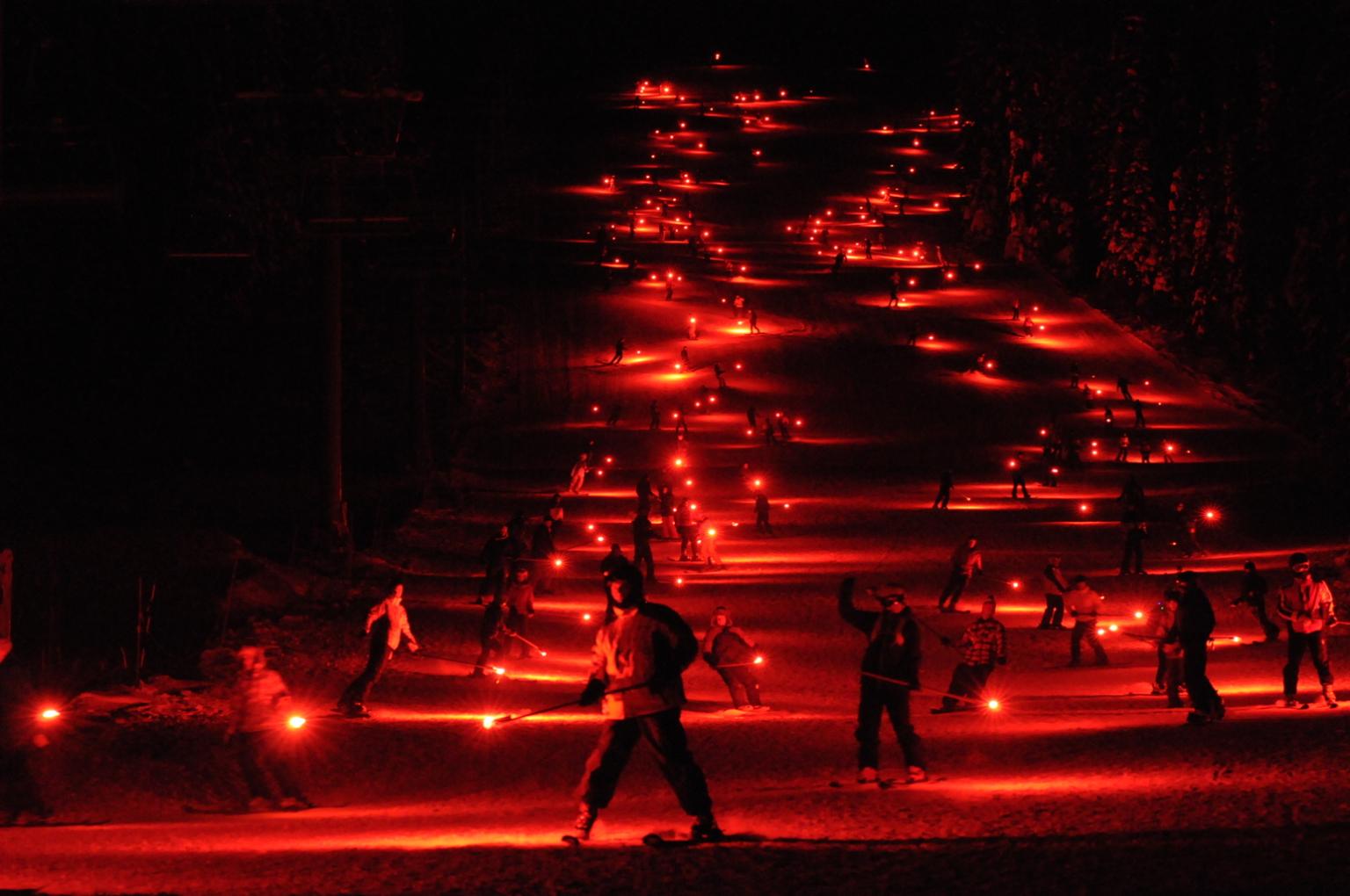 Dozens of skiers and snowboarders riding down the ski hill at night holding red torch lights
