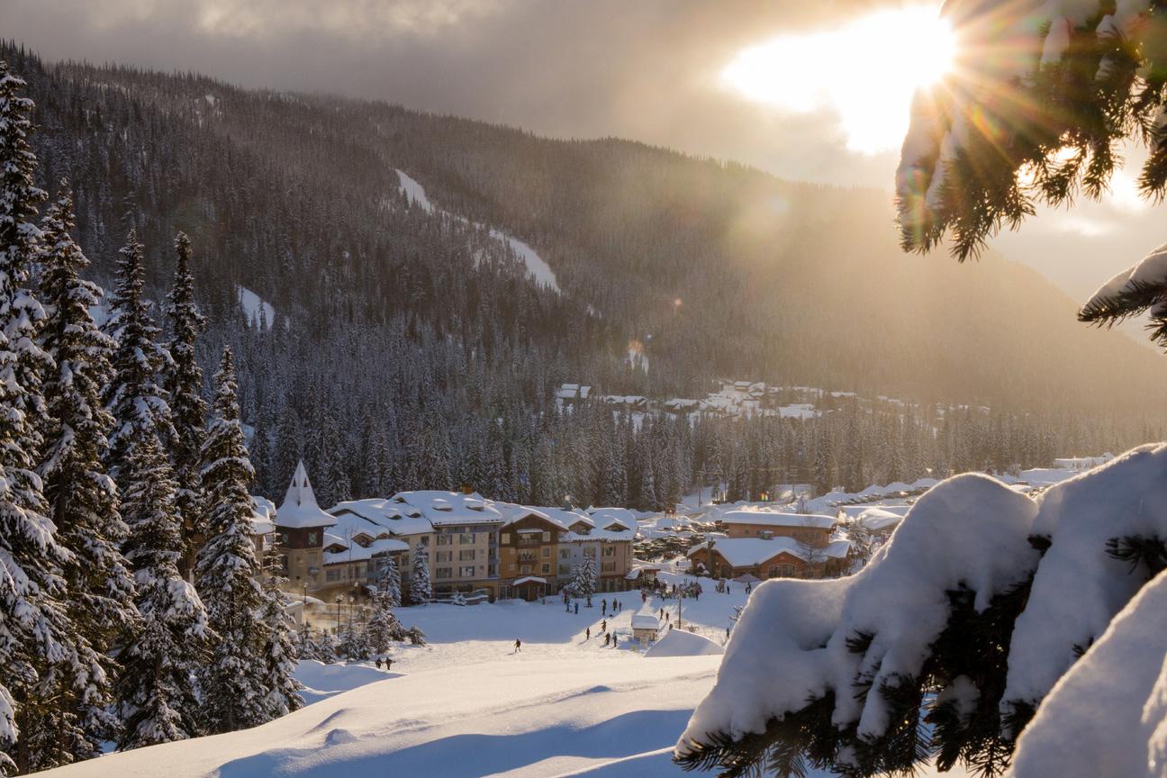 Winter view of Sun Peaks village and Mt. Morrisey taken from low on Sundance Mountain with snow covering the ground, trees, and sun peaking through overheard.