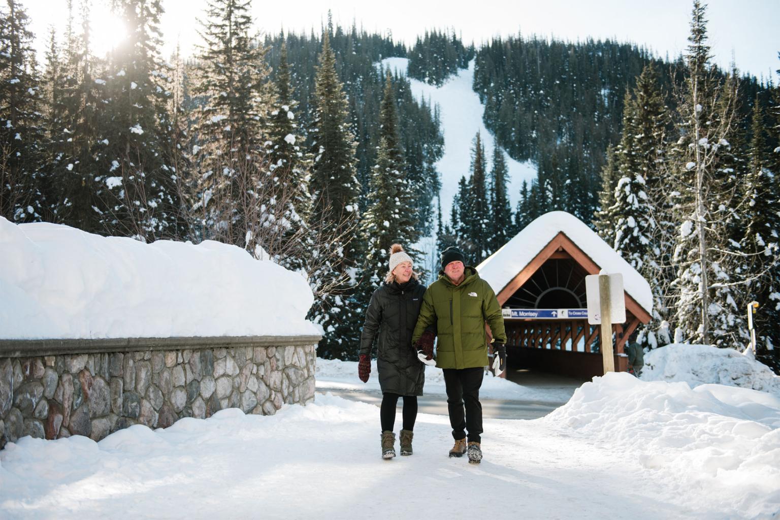 Smiling couple walking in snowy Sun Peaks village with covered bridge in the background
