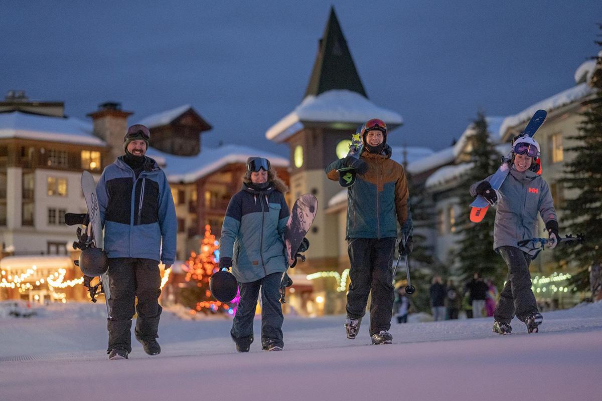 Two skiers and two boarders walk through twinkling, snowy Sun Peaks village at night.
