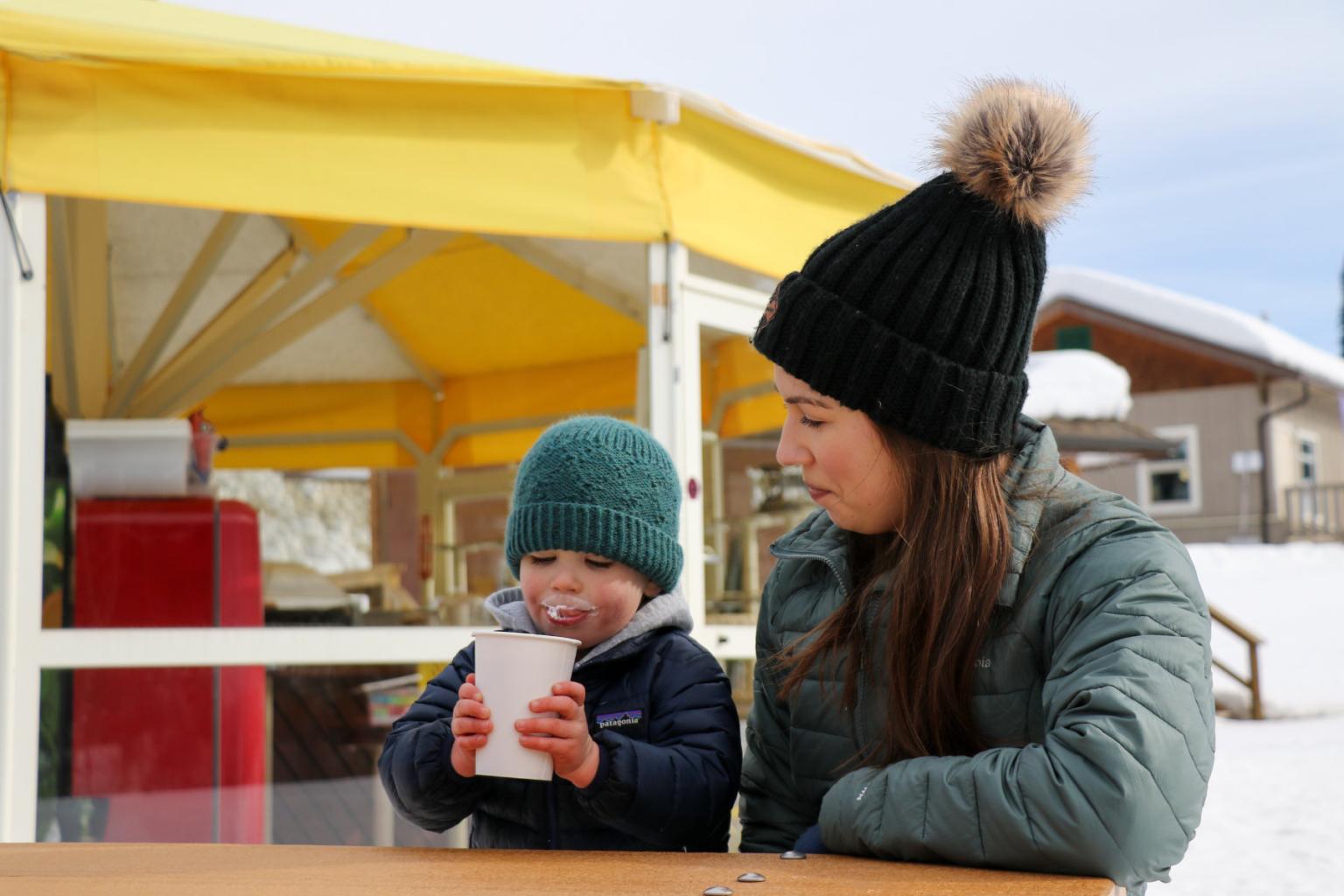 A child and mother sit at a picnic table in front of a yellow tent drinking hot chocolate