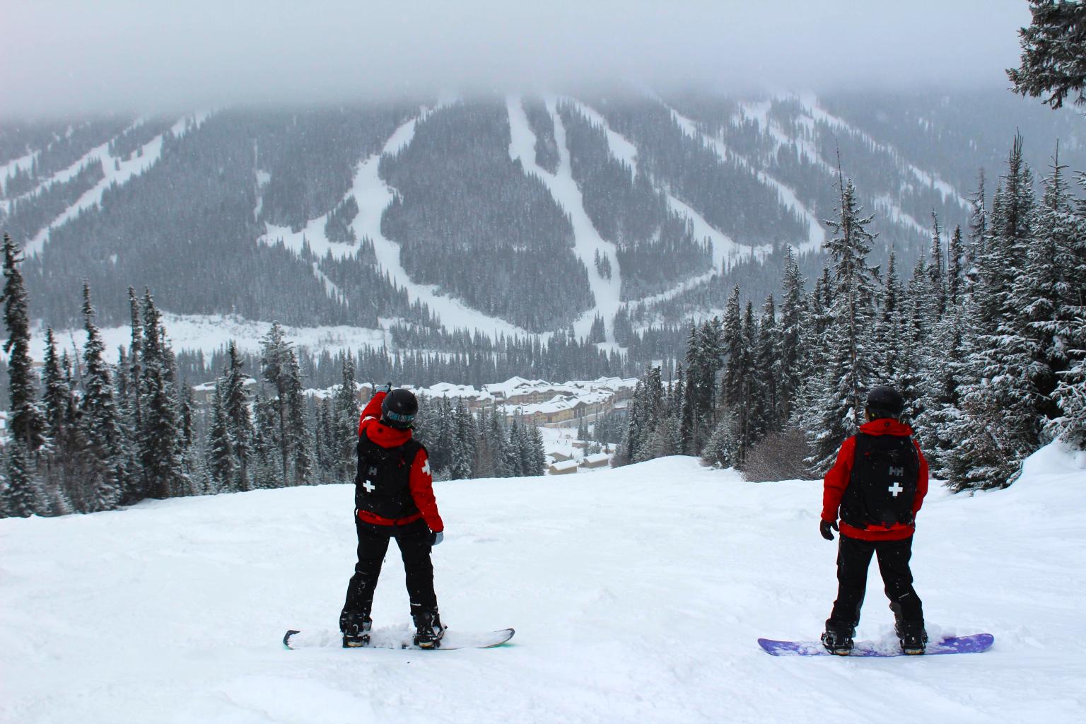 Two snowboarders in red jackets on mountain looking at a snowy mountain slope with trees and ski trails.