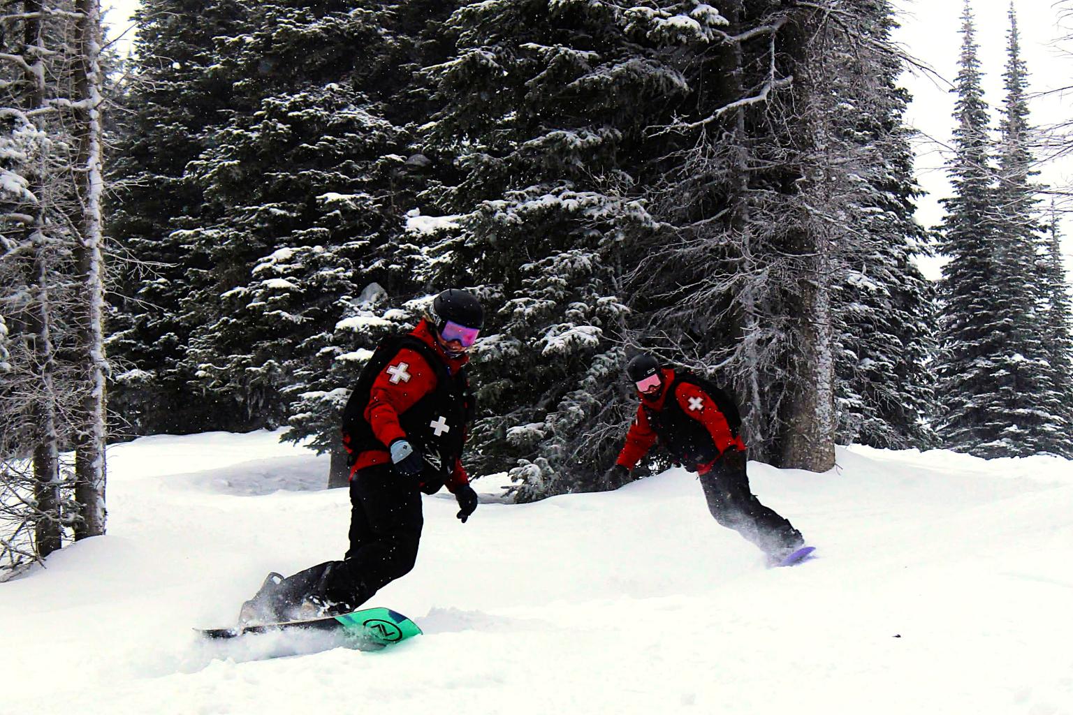 Two snowboarders in red jackets ride through snowy forest