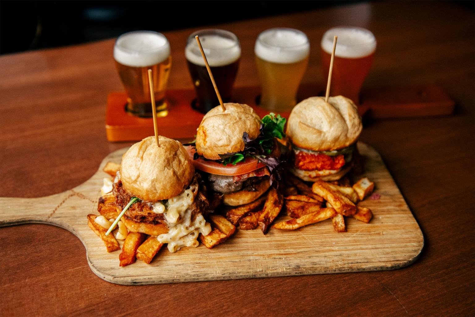 Three sliders with fries on a wooden board, flight of beer in the background.