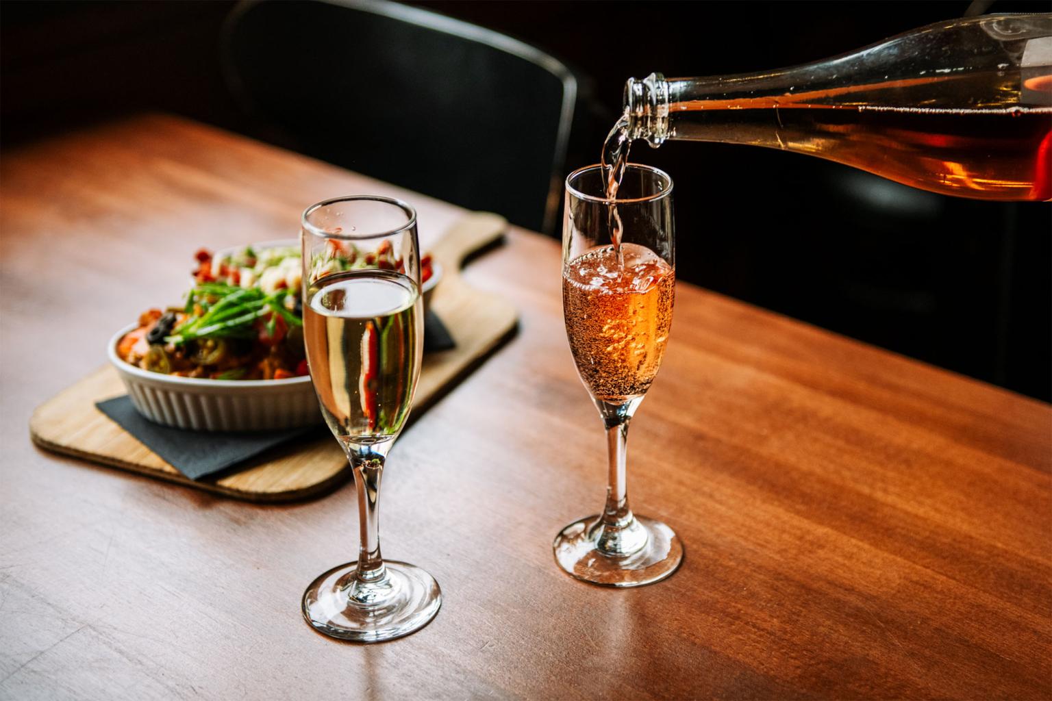 Two glasses of champagne, one being filled; loaded fries on wooden table.