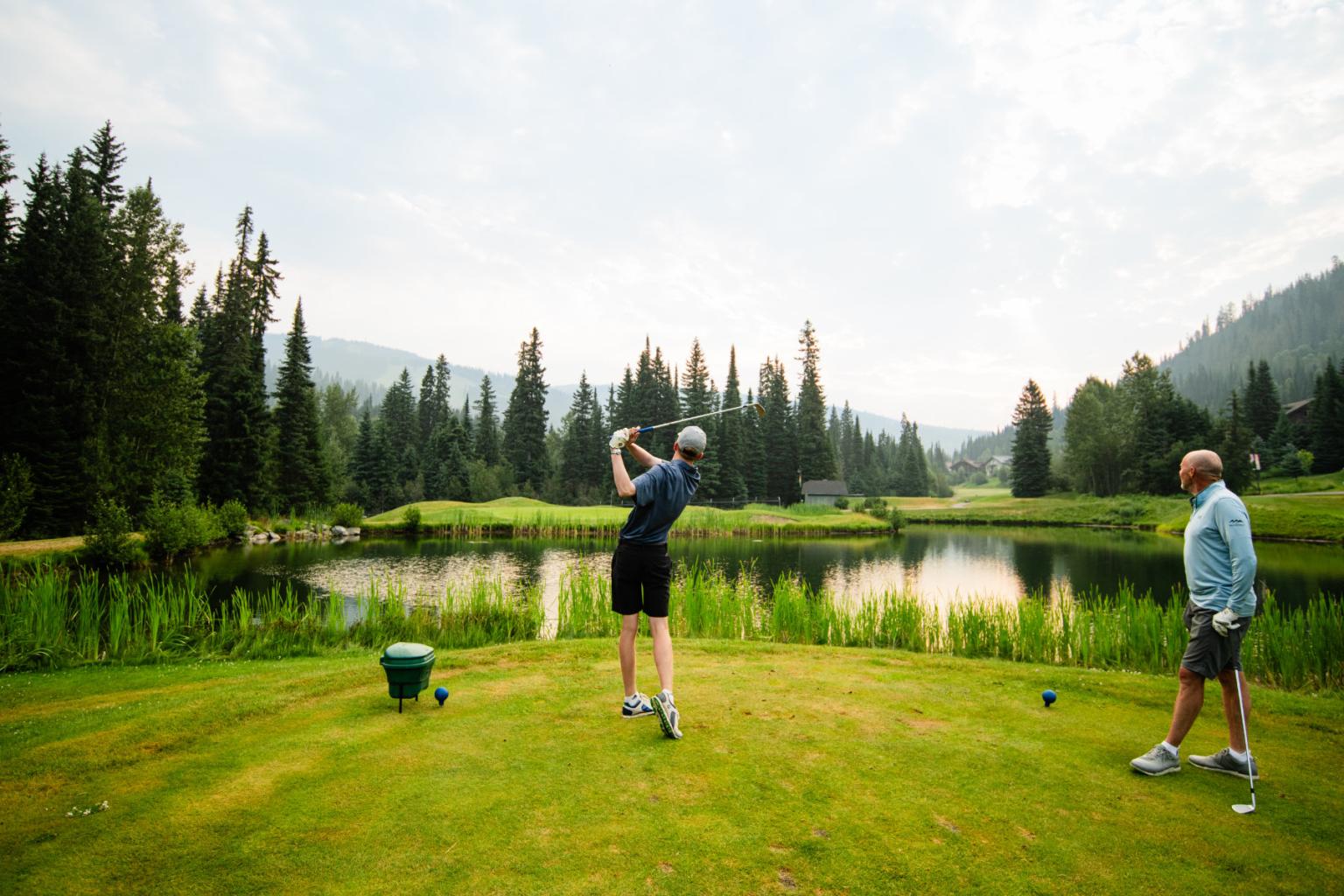 Golfer swings on a lush course by a lake, with mountains in the background.