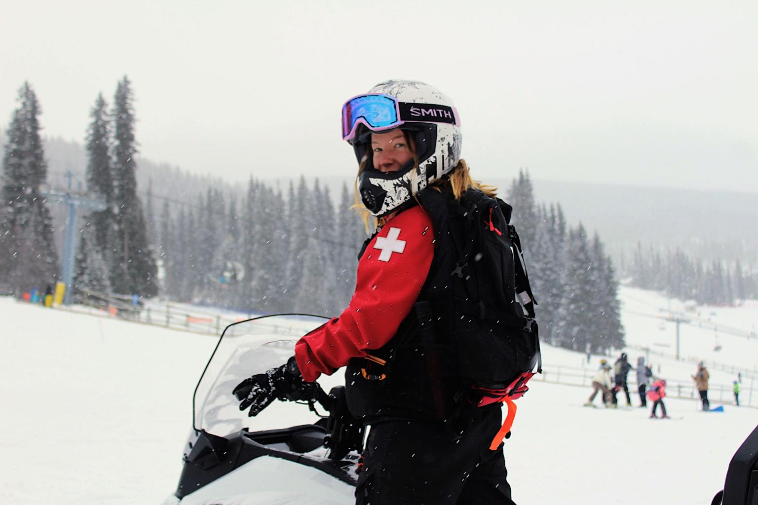 Ski patroller climbing onto snowmobile, snowy background.