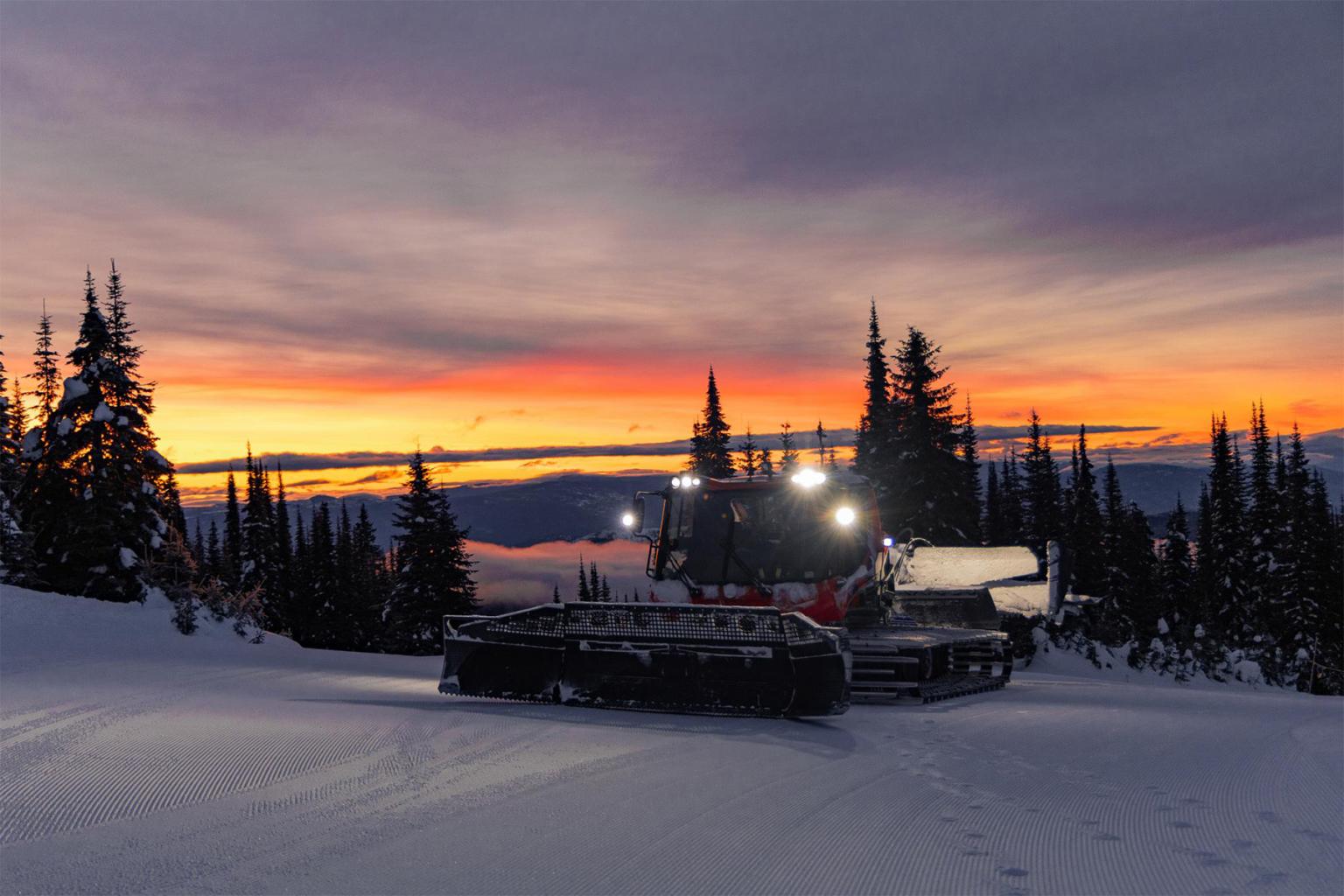 Snowcat grooming ski slope at sunset with colorful sky.