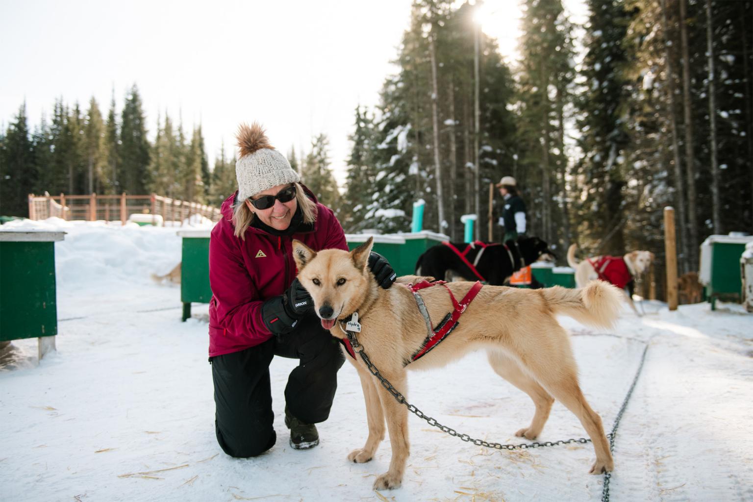 Woman in a red jacket in snow petting a sled dog near trees.