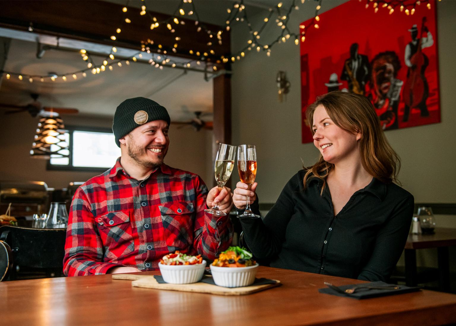 Emily and Ilkay toasting with champagne at a cozy restaurant table, with string lights above.