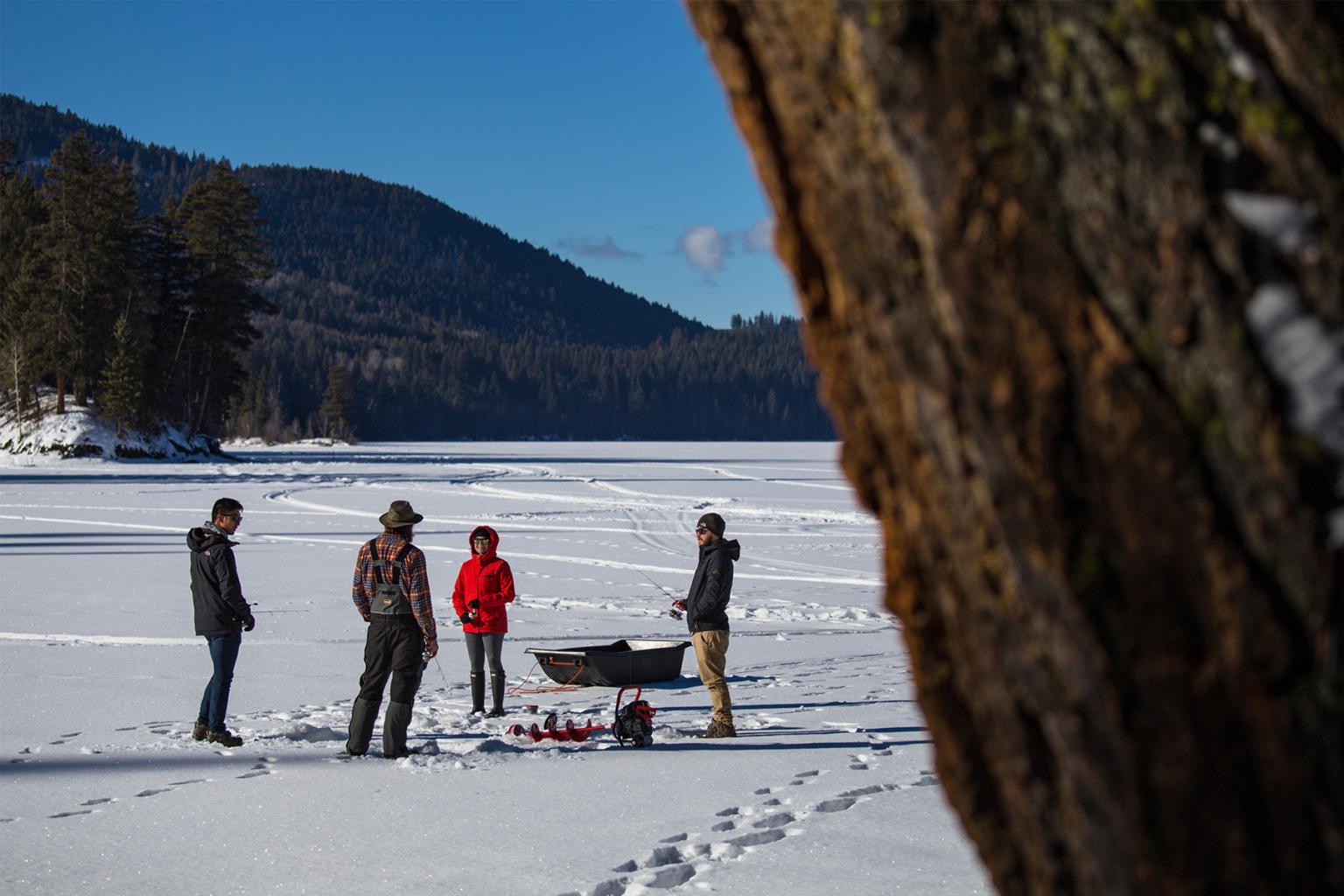 Four people stand on a frozen lake to ice fish, snow-covered hills in the background.