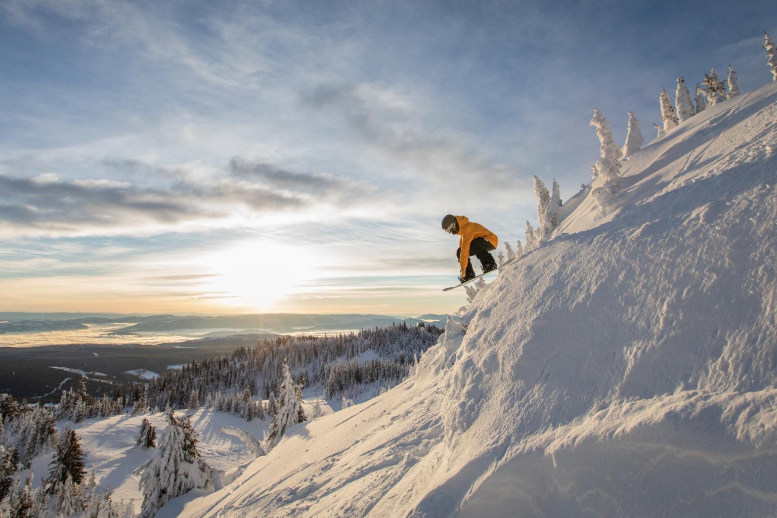 Snowboarder in yellow descends a snowy mountain slope at sunset.