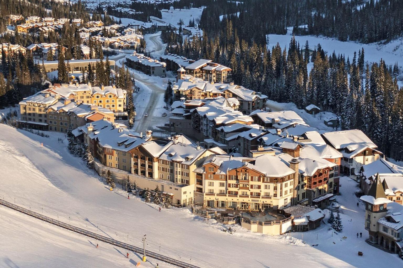 Snow-covered village with chalet-style buildings, surrounded by pine forest.