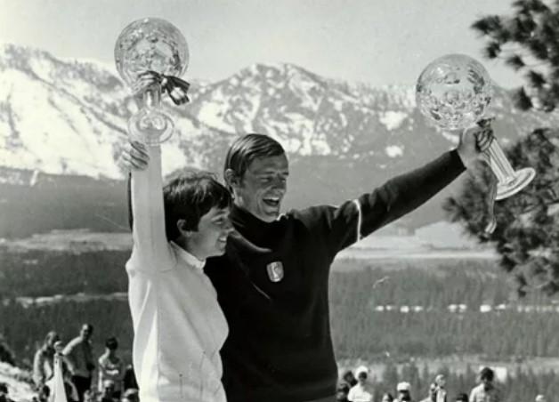 Two people smiling(Nancy is one), holding trophies with snowy mountains in the background.