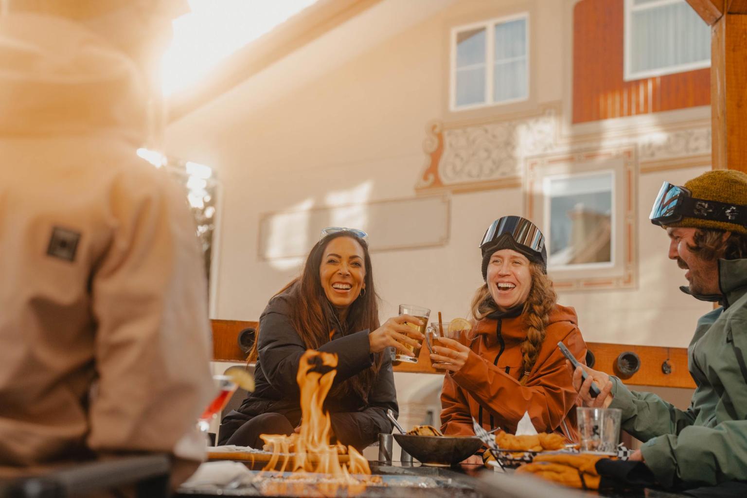 Group of people in winter attire smiling around a fire table with lunch and drinks.
