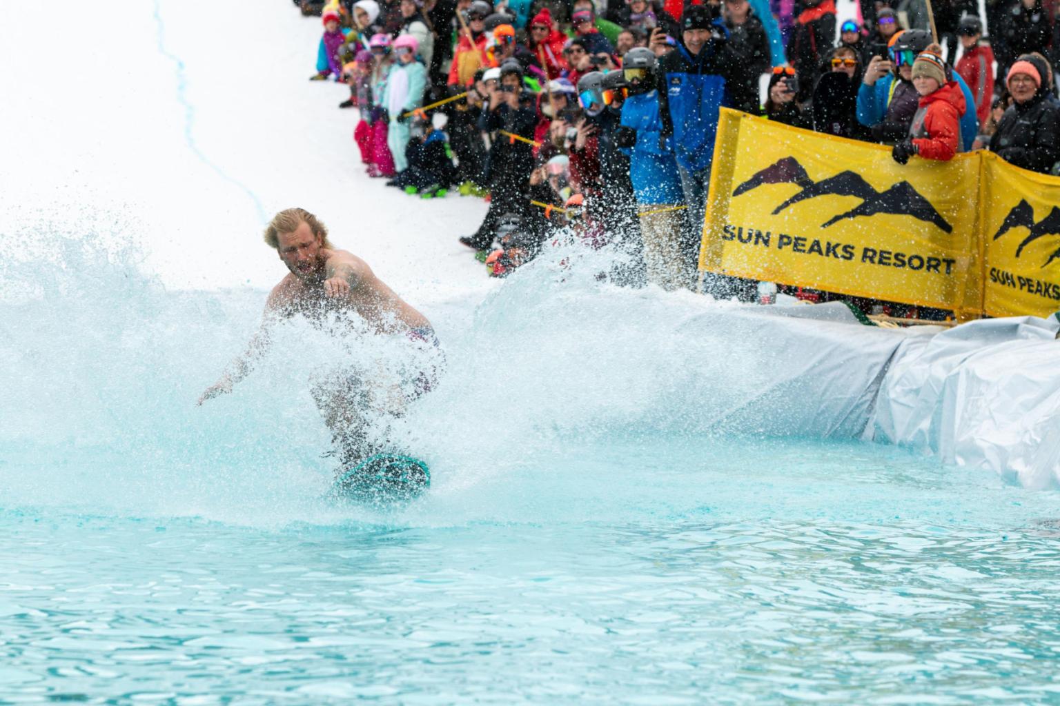 Snowboarder splashes through water at event; crowd watches, bright banners in background.