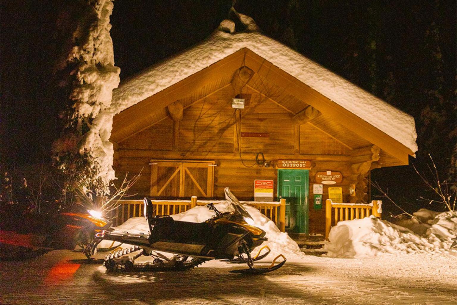 Snow-covered cabin at night with a snowmobile in front.