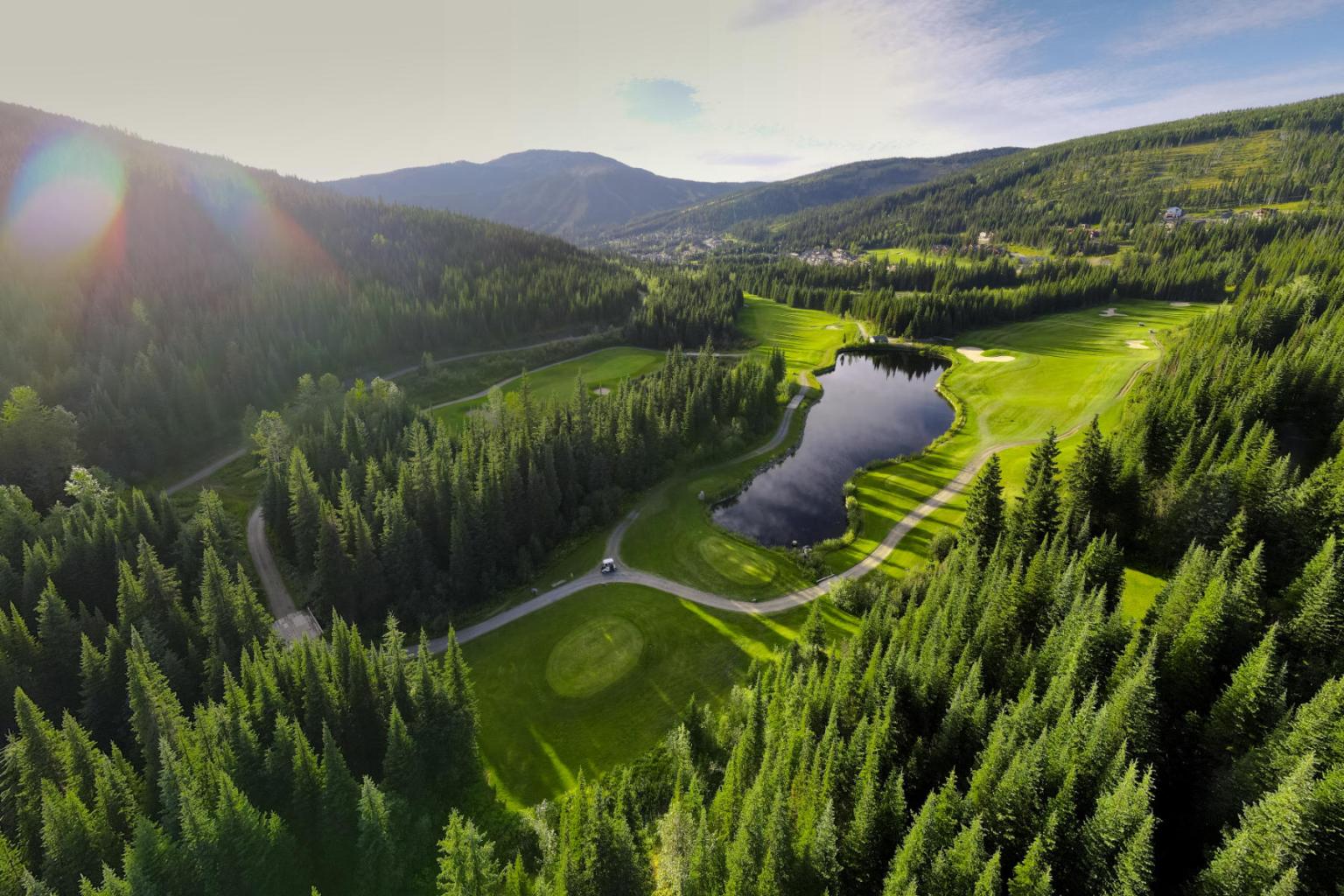 Mountainous landscape with a golf course, trees, and a small pond under a clear sky.