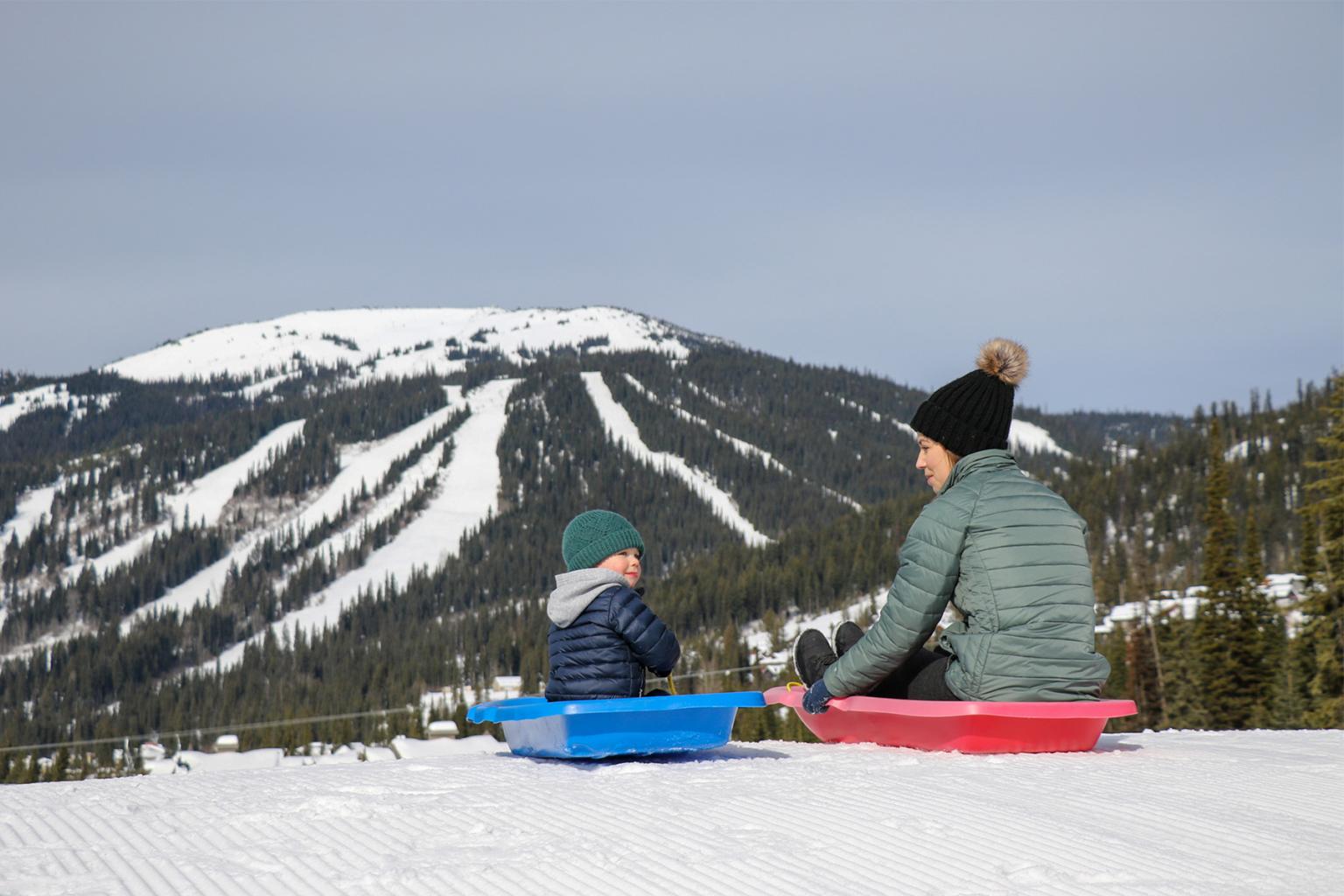 Two people sledding on snow, mountain in the background.