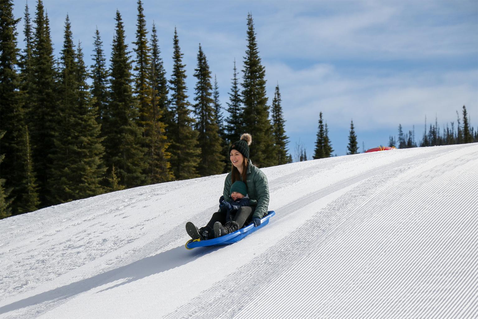 Mother and child sledding down a snowy hill, surrounded by pine trees.