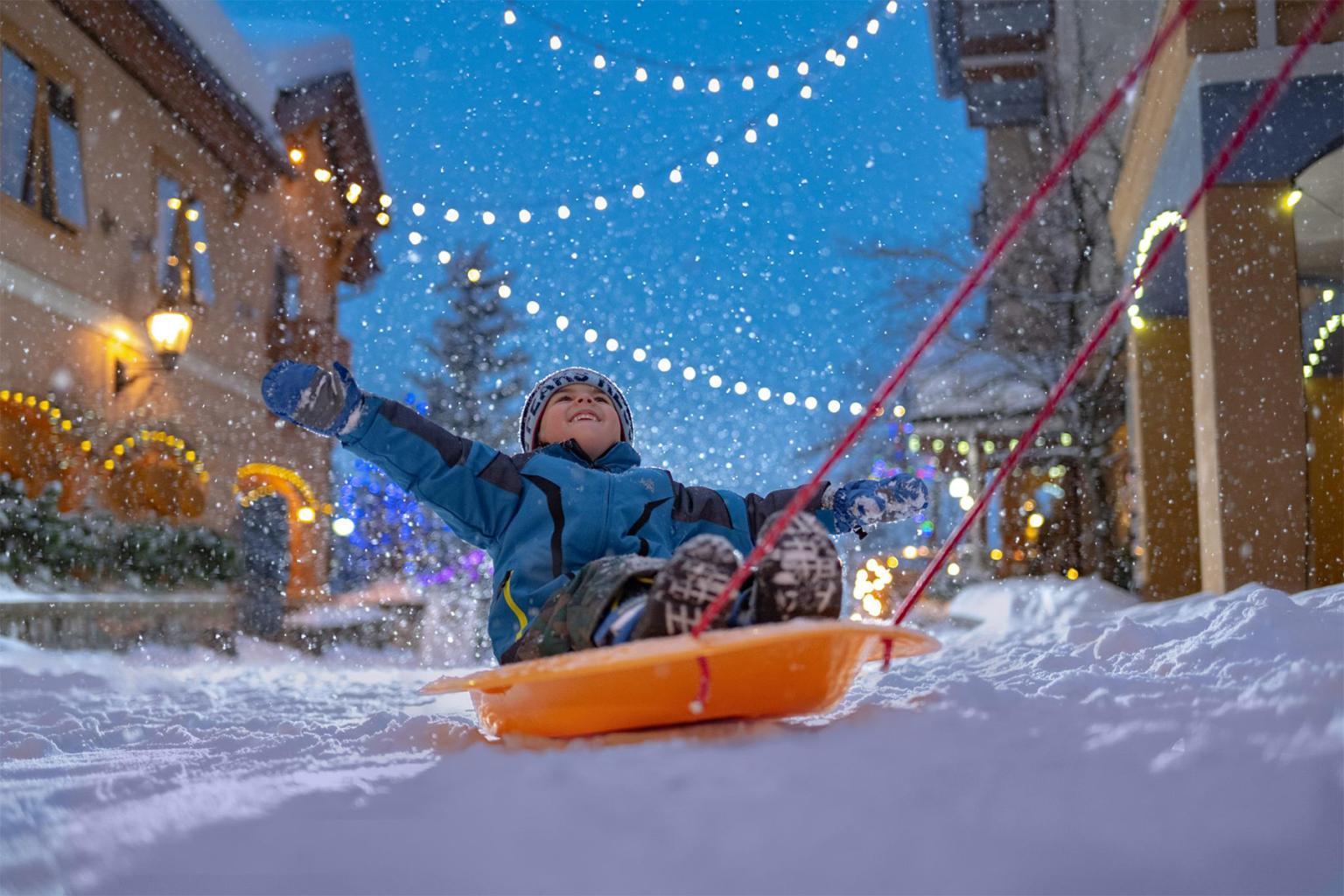 Child sledding in snowy village, joyful expression, string lights overhead.