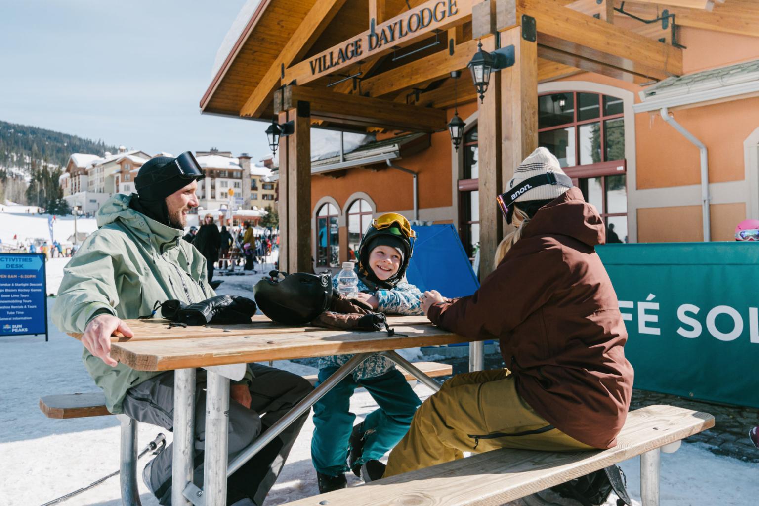 Three people in ski gear sitting at a snowy outdoor table, smiling.