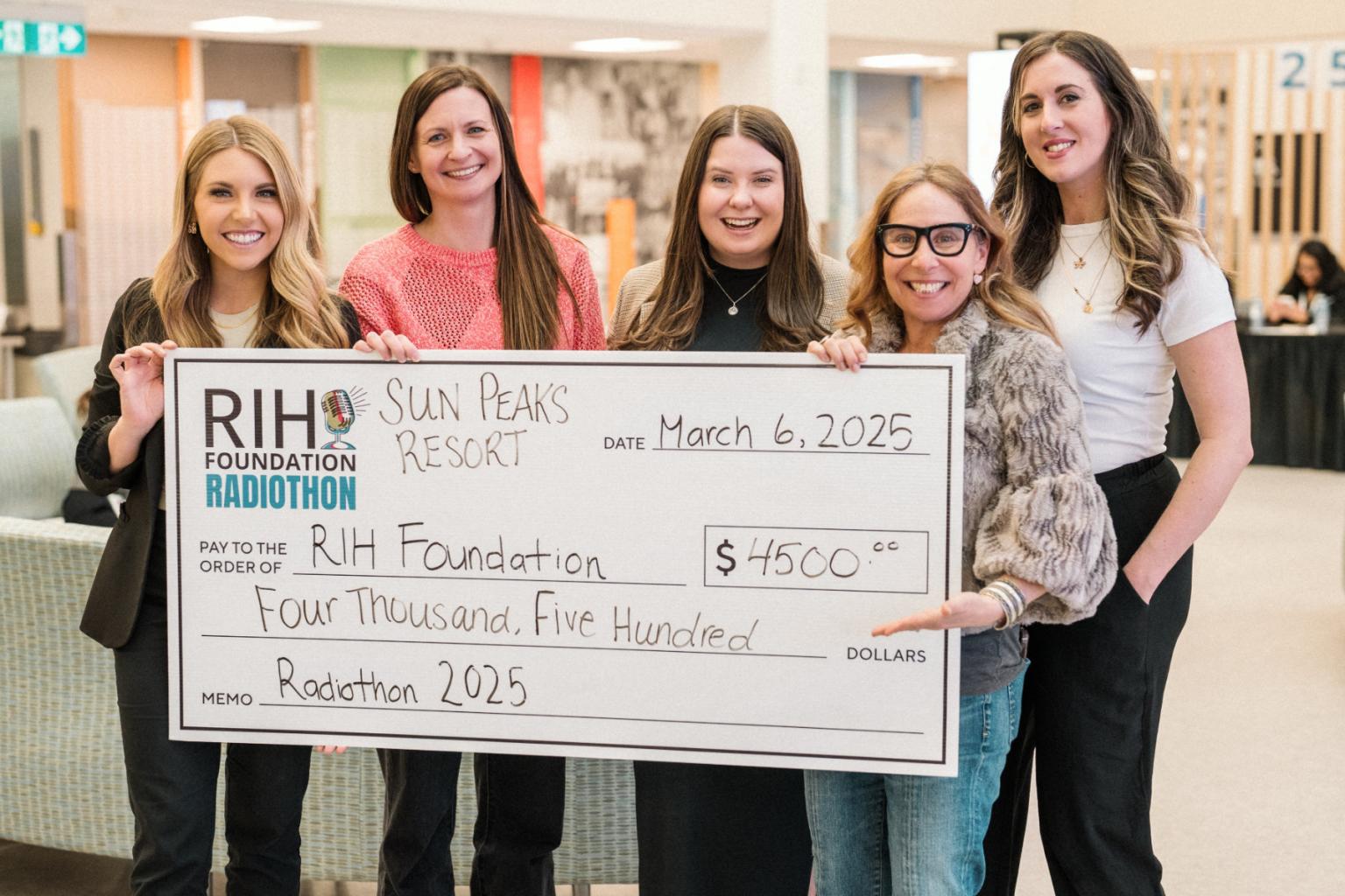 A group of five women smiling, holding a large check indoors.