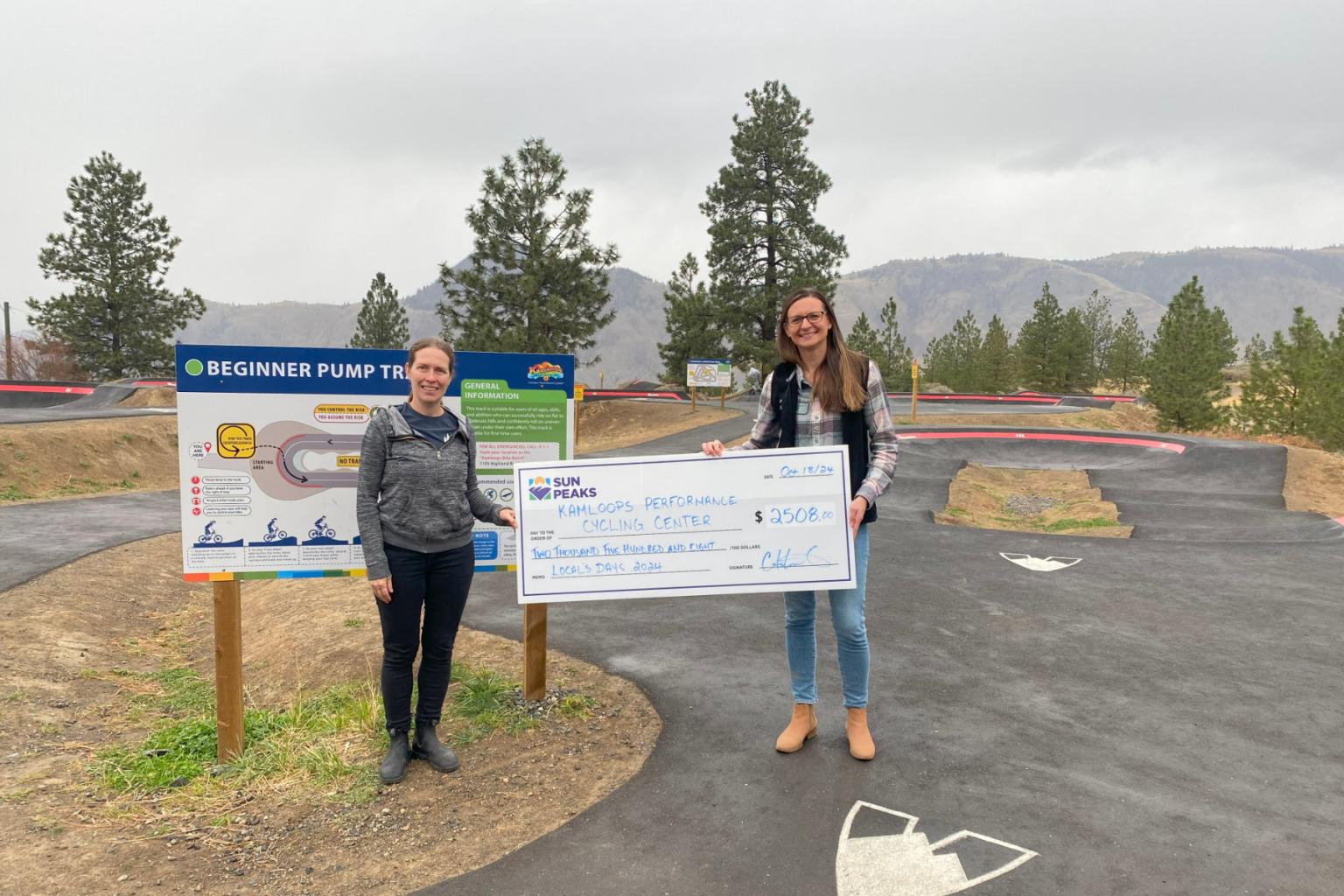 Two women hold a large check at an outdoor park with trees and mountains.