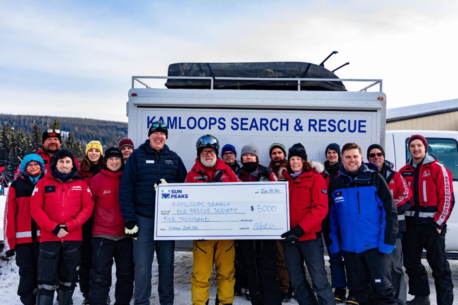 Rescue team in winter gear holding a large check, standing by a truck.