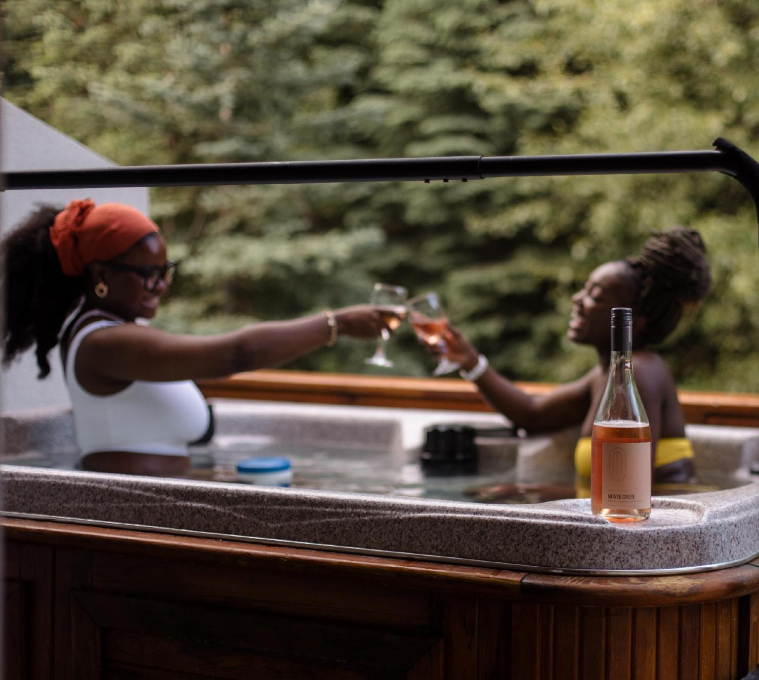 Two women clink glasses in a hot tub with a forest backdrop.