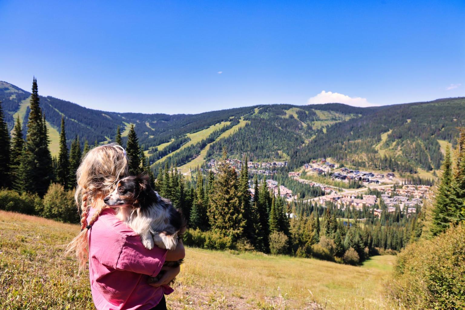 Woman holding a dog, overlooking a scenic valley with mountains and village.