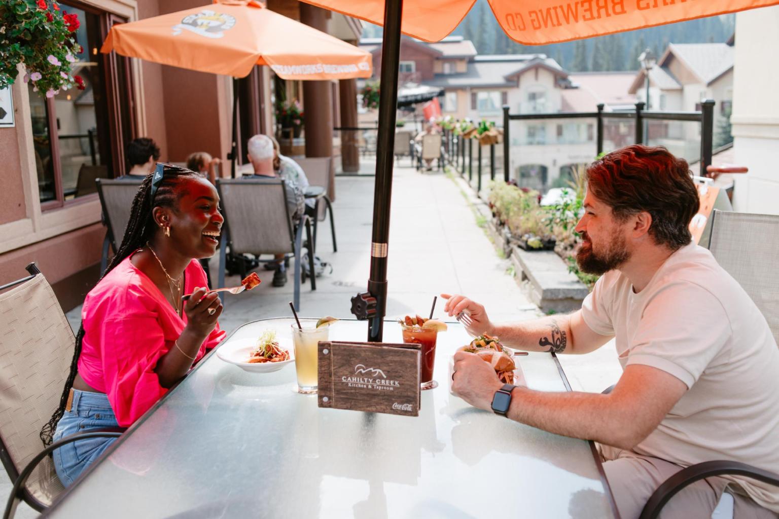Two people dining outdoors under orange umbrellas, enjoying a sunny day.