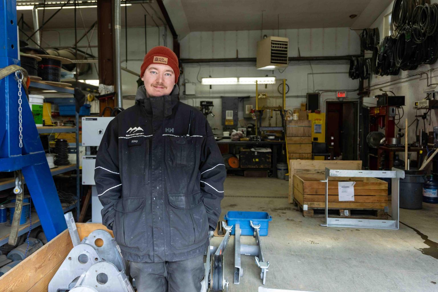 Man in a workshop wears a red hat and black jacket, surrounded by tools and machinery.
