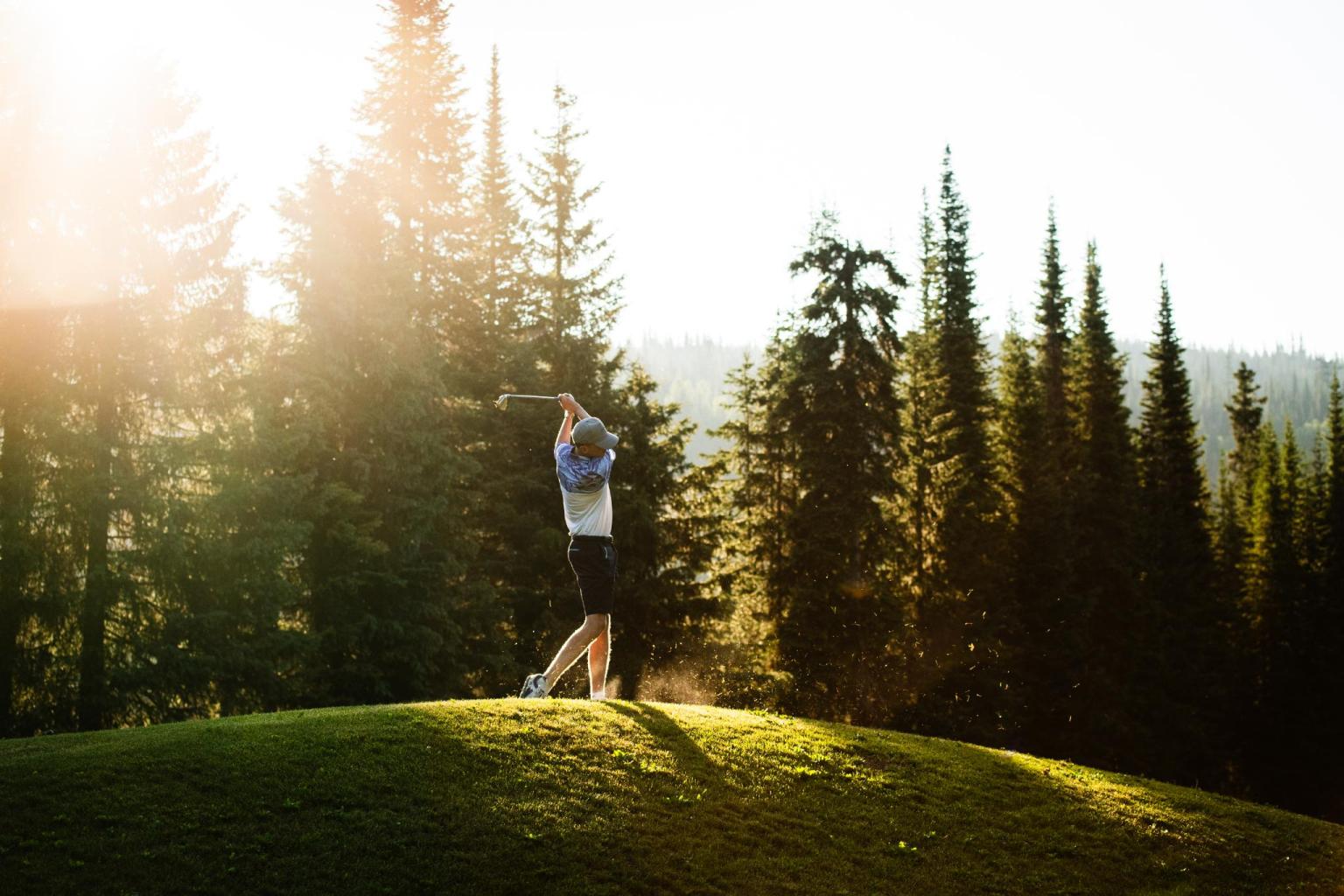 Golfer swinging on the sunny Sun Peaks Golf Course, surrounded by tall trees.