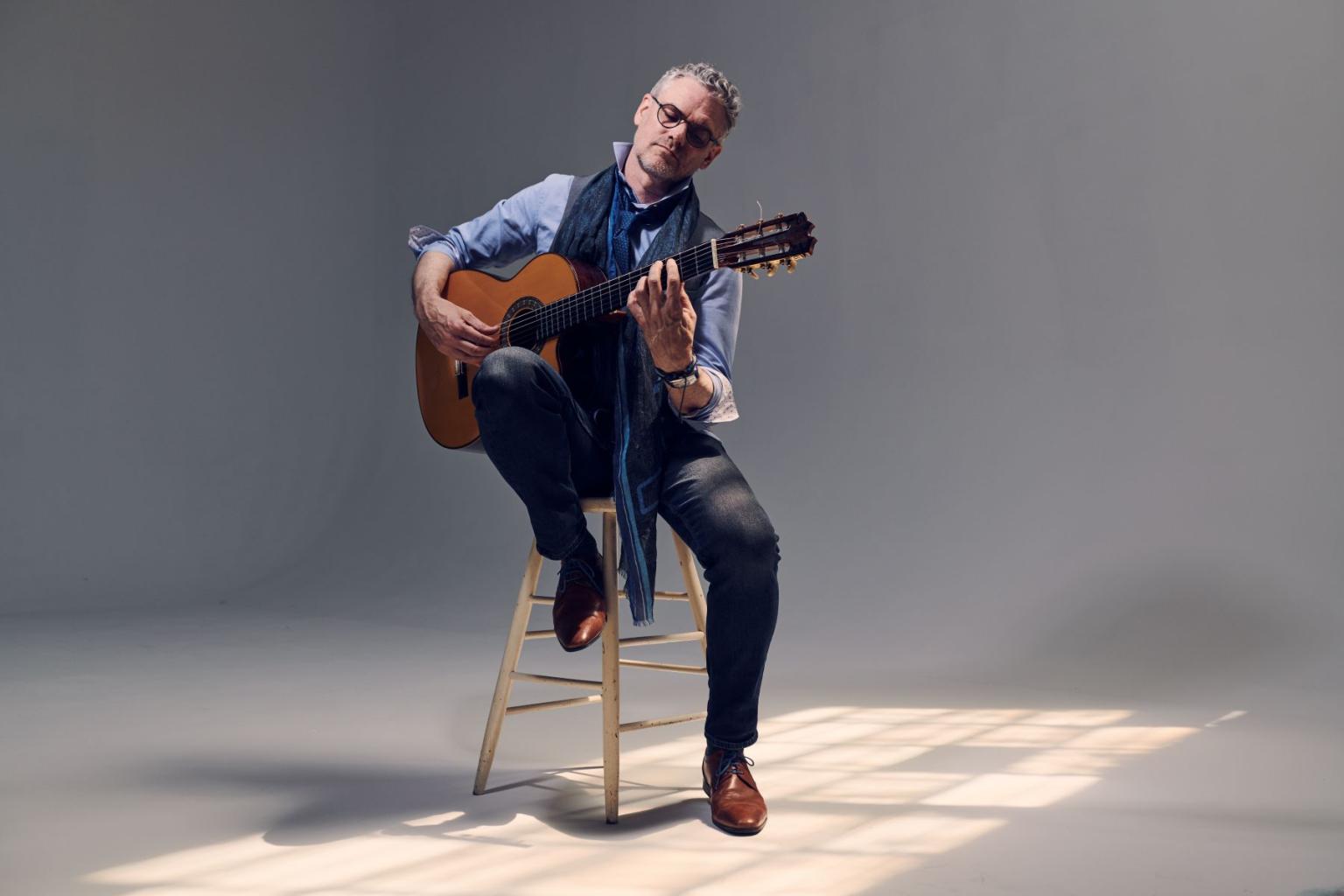 Man playing guitar on a stool in a softly lit studio.