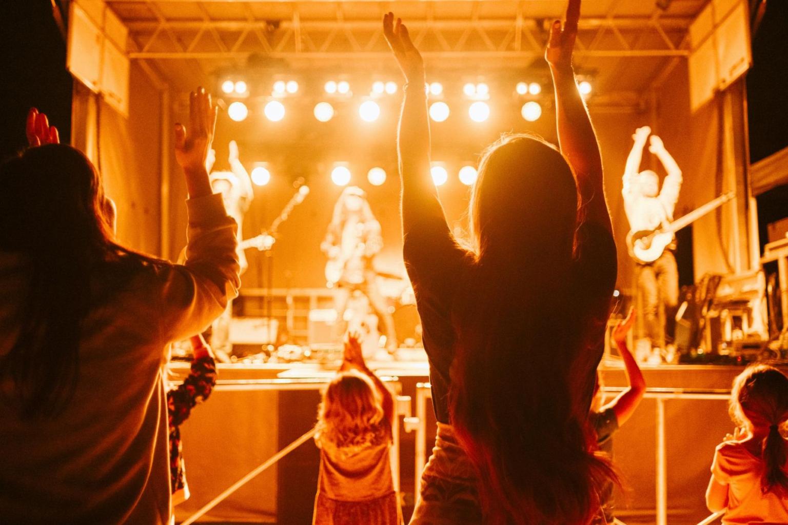 Concert audience cheering as band performs on stage with bright lights.