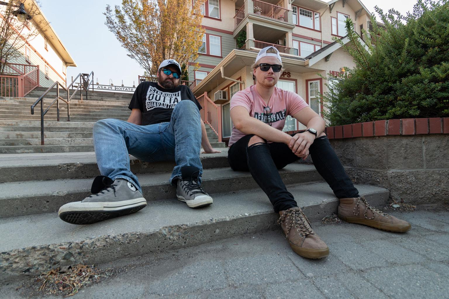 Two men sit casually on outdoor steps, with a building and trees in the background.