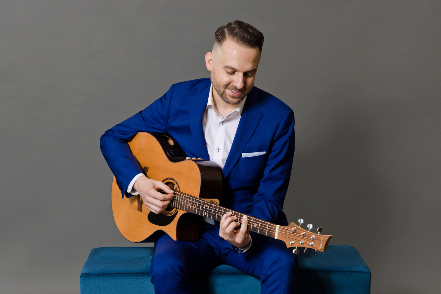 Man in blue suit playing acoustic guitar, smiling, seated on a blue bench.